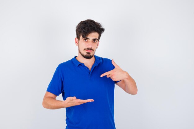 Young man stretching one hand as holding something and pointing to it with index finger in blue t-shirt and looking serious