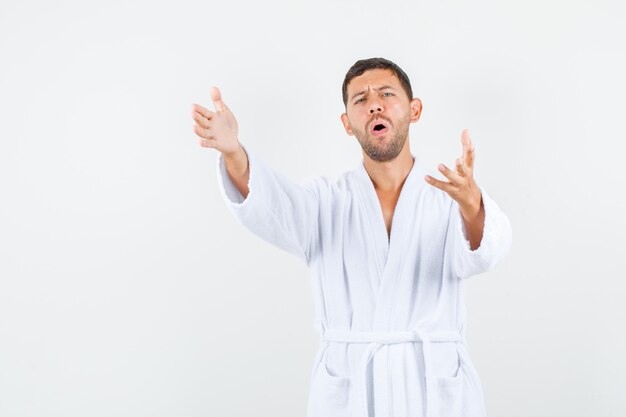 Young man stretching out hands in white bathrobe front view.