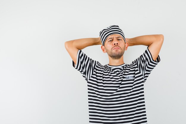 Young man in striped t-shirt hat holding hands behind head and looking pensive  