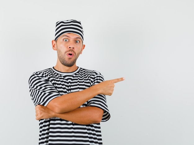 Young man in striped t-shirt hat pointing to the side and looking surprised  