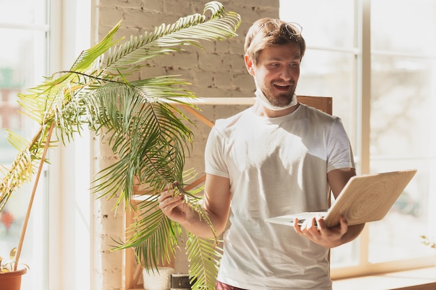 Young man studying at home during online courses for gardener, biologist, florist. Getting profession while isolated, quarantine against coronavirus spreading. Using laptop, smartphone, headphones.