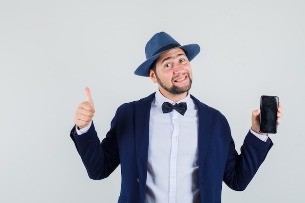 Young man in suit, hat holding mobile phone with thumb up and looking cheery , front view.