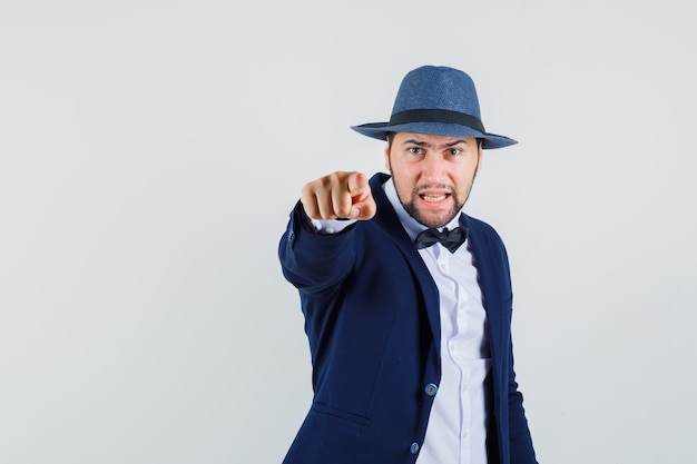 Young man in suit, hat pointing at camera and looking furious , front view.