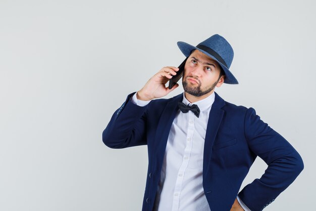 Young man in suit, hat talking on mobile phone and looking irresolute , front view.