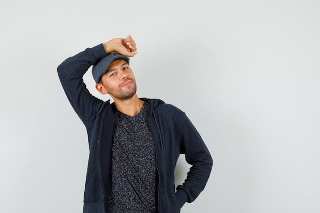 Free photo young man in t-shirt, jacket, cap holding raised arm over head and looking handsome , front view.