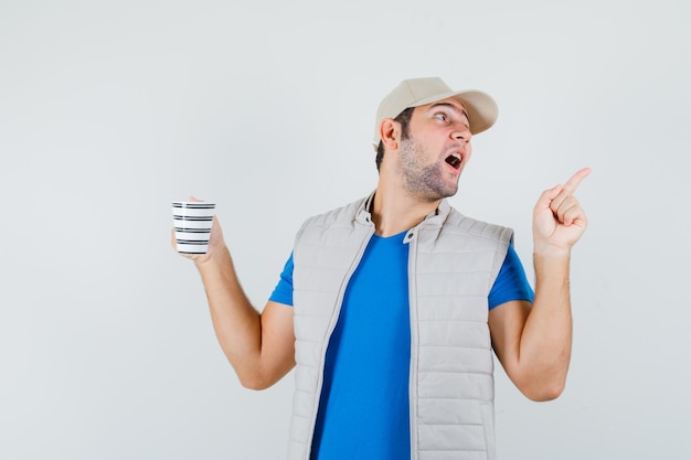 Young man in t-shirt, jacket, cap pointing up, holding cup of drink and looking amazed , front view.