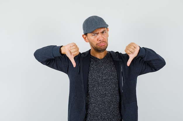 Free photo young man in t-shirt, jacket, cap showing double thumbs down and looking disappointed , front view.