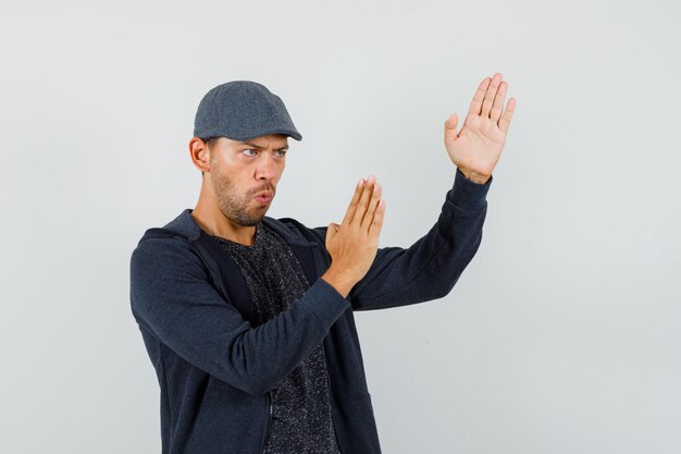 Young man in t-shirt, jacket, cap showing karate chop gesture and looking confident , front view.