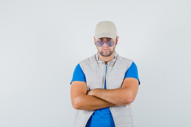 Free photo young man in t-shirt, jacket, cap standing with crossed arms and looking confident , front view.