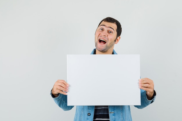 Young man in t-shirt, jacket holding blank canvas and looking glad