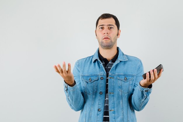 Young man in t-shirt, jacket holding calculator and looking confused