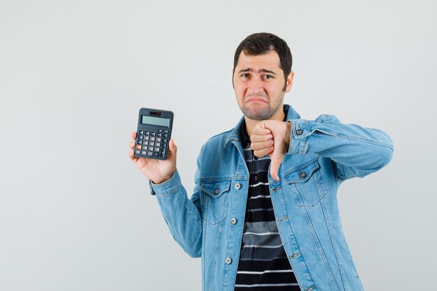 Young man in t-shirt, jacket holding calculator, showing thumb down and looking disappointed