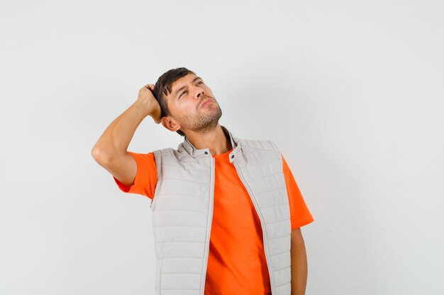 Young man in t-shirt, jacket scratching head while looking up and looking pensive , front view.