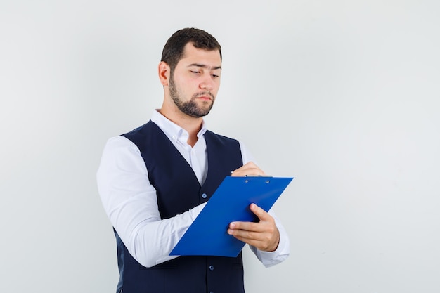 Young man taking notes on clipboard in shirt and vest and looking busy