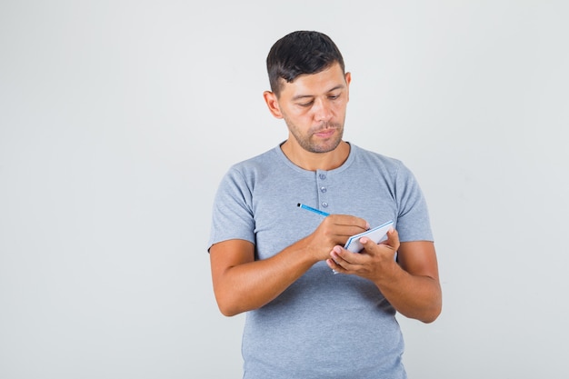 Young man taking notes on notebook in grey t-shirt and looking careful.