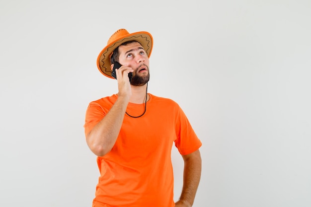 Young man talking on mobile phone in orange t-shirt, hat and looking pensive. front view.