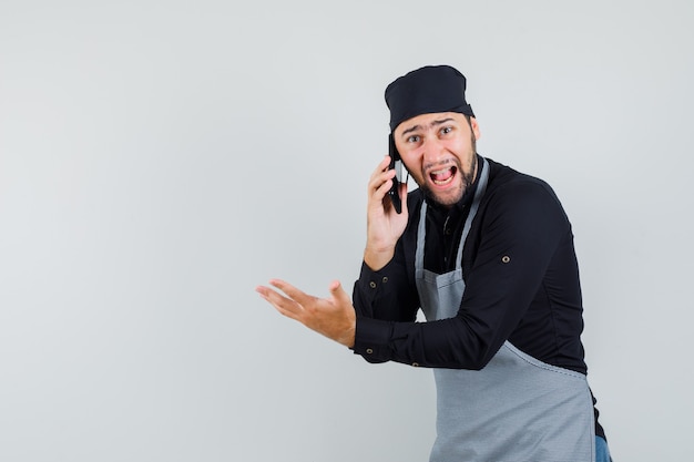 Young man talking on mobile phone in shirt, apron and looking furious , front view.