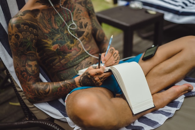 Free photo a young man in tattoos wearing headphones listens to music and draws in a notebook.