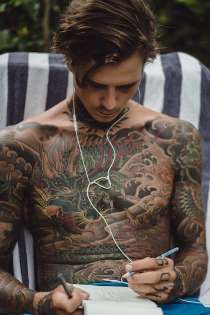 Free Photo a young man in tattoos wearing headphones listens to music and draws in a notebook.