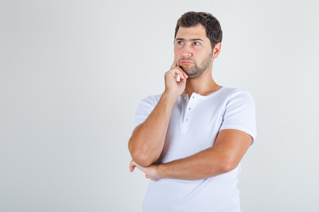 Young man thinking with finger on cheek in white t-shirt.