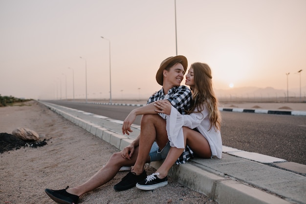 Young man in trendy hat looking with love at his graceful girlfriend in white shirt, while resting after walk. Couple of travelers sitting near to road and gently embracing with sunset