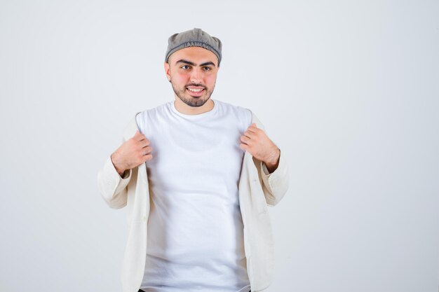 Young man trying to take off jacket and posing at front in white t-shirt, jacket and gray cap and looking happy