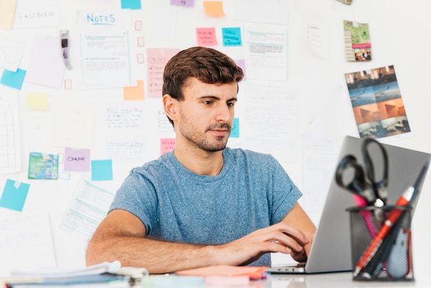 Free photo young man typing on laptop against wall with notes