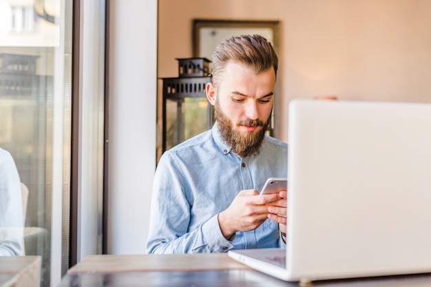 Free photo young man using cellphone with laptop on desk