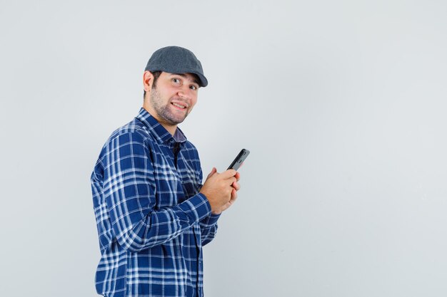 Young man using mobile phone in shirt, cap and looking cheerful , front view.