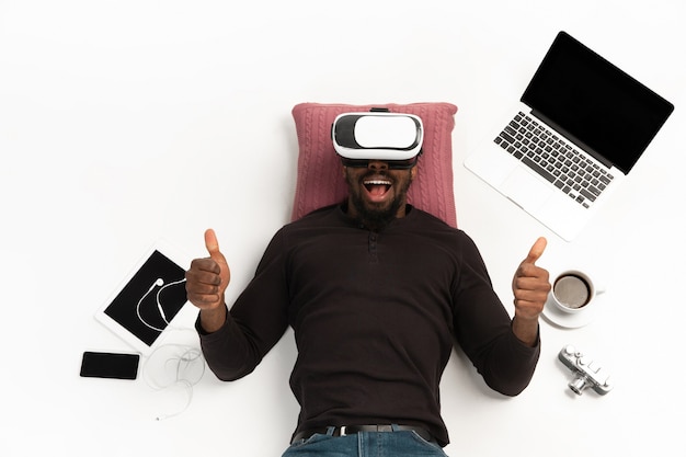 Young man using vr headset surrounded by gadgets isolated on white floor