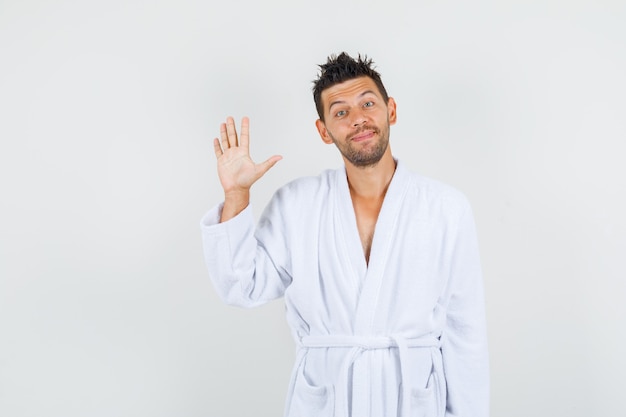 Free photo young man waving hand for greeting in white bathrobe and looking cheerful. front view.