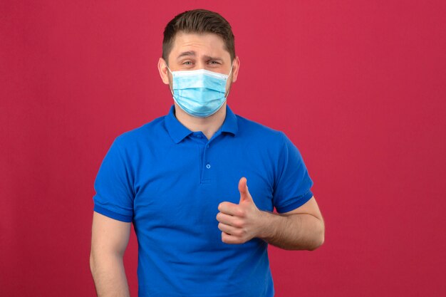 Young man wearing blue polo shirt in medical protective mask smiling cheerfully showing thumb up over isolated pink wall