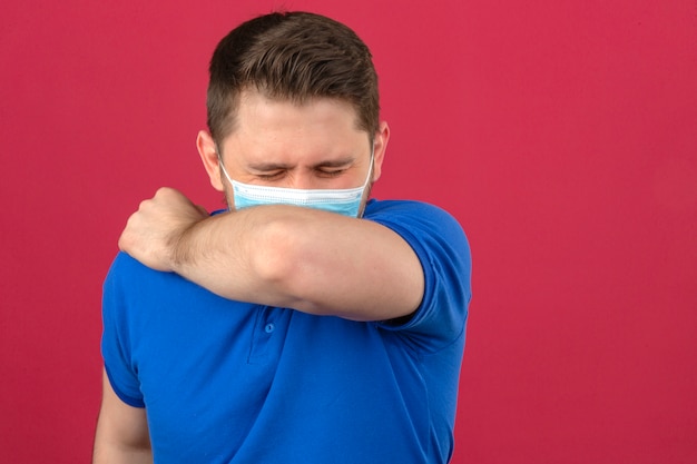 Young man wearing blue polo shirt in medical protective mask sneezing coughing into his arm or elbow to prevent spread covid-19coronavirus over isolated pink wall