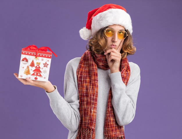 Young man wearing christmas santa hat and yellow glasses with warm scarf around his neck holding christmas gift  looking up with pensive expression thinking  standing over purple  wall