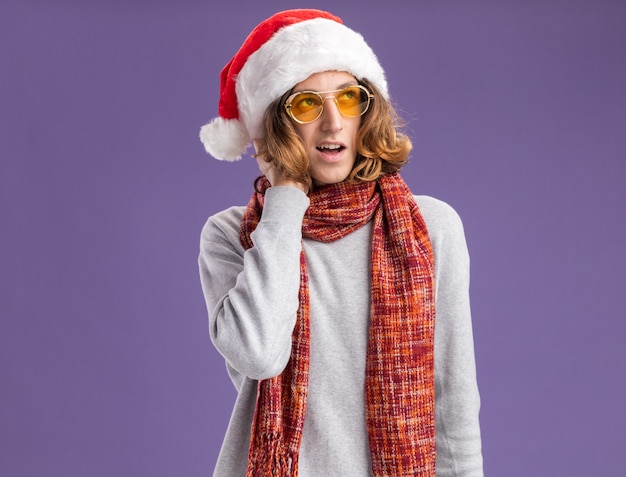 Free Photo young man wearing christmas santa hat and yellow glasses with warm scarf around his neck looking up with pensive expression thinking positive standing over purple  wall