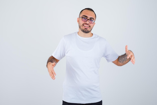 Young man wearing glasses and stretching hands as holding something in white t-shirt and black pants and looking happy