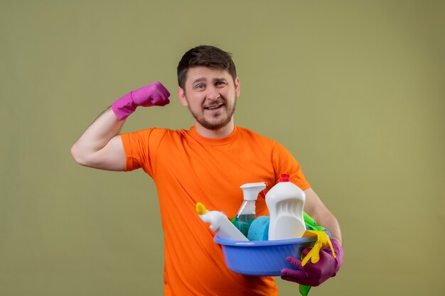 Young man wearing orange t-shirt and rubber gloves holding basin with cleaning tolls smiling showing biceps ready to clean concept standing over green backgroun