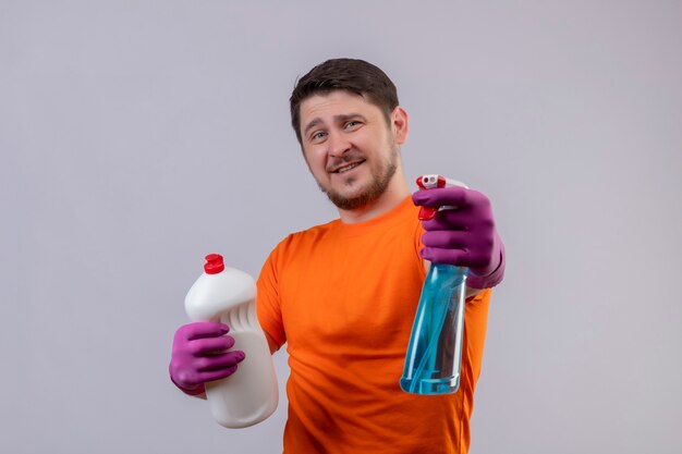 Free photo young man wearing orange t-shirt and rubber gloves holding bottles with cleaning supplies smiling cheerfully