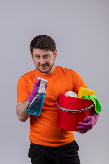 Young man wearing orange t-shirt and rubber gloves holding bucket with cleaning tools and cleaning spray