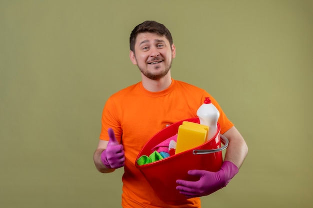 Young man wearing orange t-shirt and rubber gloves holding bucket with cleaning tools smiling cheerfully positive and happy showing thumbs up standing over green wall 2