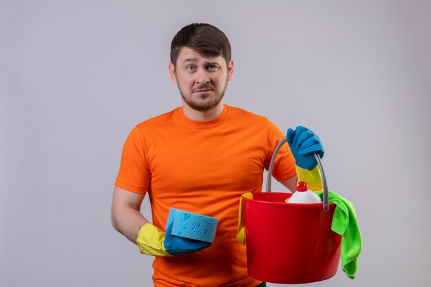 Young man wearing orange t-shirt and rubber gloves holding bucket with cleaning tools and sponge displeased and very anxious standing over white wall