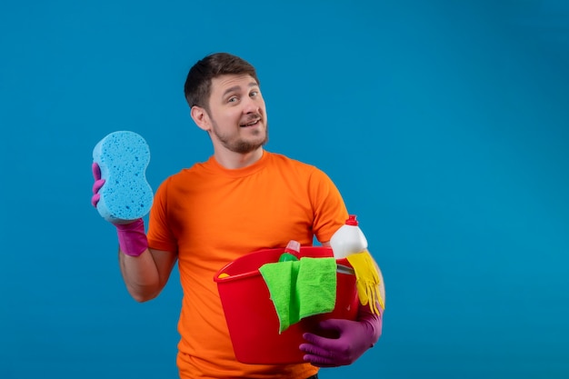 Young man wearing orange t-shirt and rubber gloves holding bucket with cleaning tools and sponge