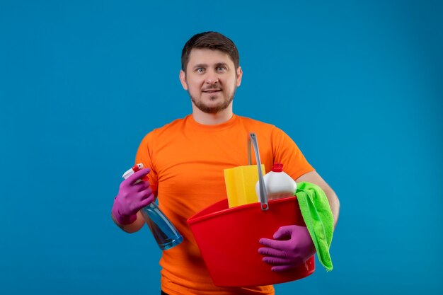 Young man wearing orange t-shirt and rubber gloves holding bucket with cleaning tools