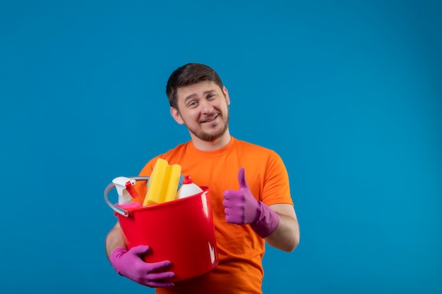 Free photo young man wearing orange t-shirt and rubber gloves holding bucket with cleaning tools