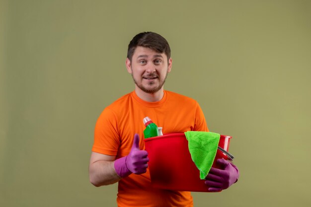 Young man wearing orange t-shirt and rubber gloves holding bucket with cleaning tools