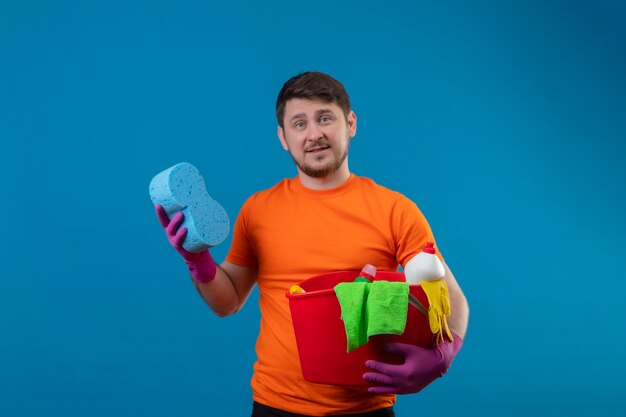 Young man wearing orange t-shirt and rubber gloves holding bucket with cleaning tools