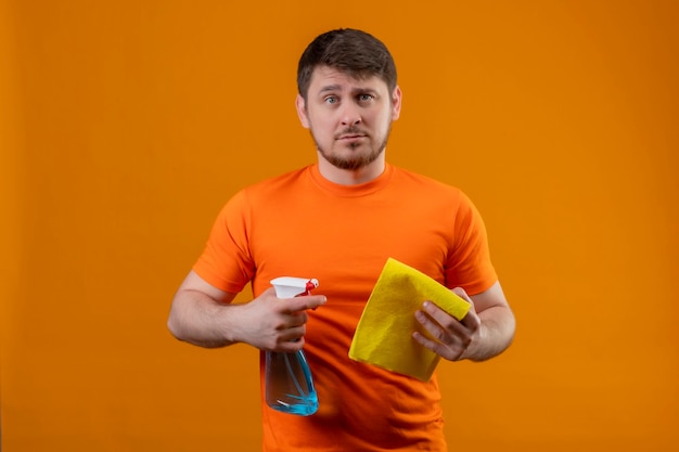 Young man wearing orange t-shirt and rubber gloves holding cleaning spray and rug displeased with sad expression