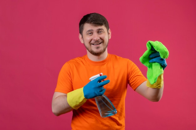 Young man wearing orange t-shirt and rubber gloves holding cleaning spray and rug smiling cheerfully positive and happy standing over pink wall