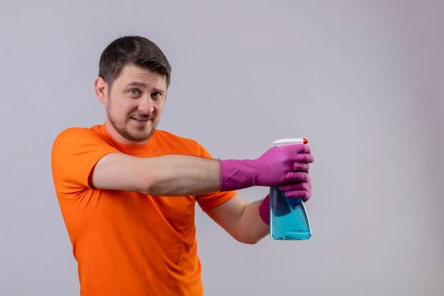 Young man wearing orange t-shirt and rubber gloves holding cleaning spray smiling using spray standing over white wall