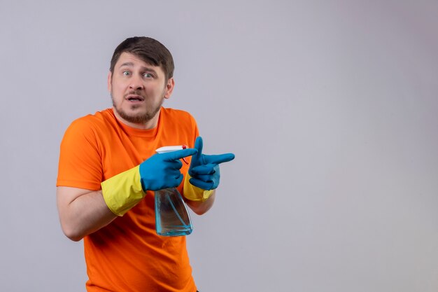 Young man wearing orange t-shirt and rubber gloves holding cleaning spray worried pointing with fingers to the side